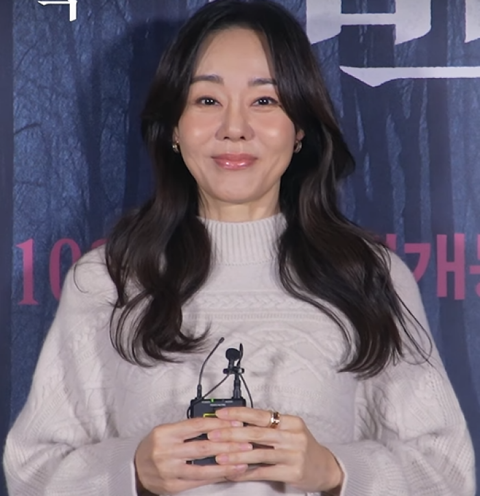 Yunjin Kim smiling at a public event, wearing a beige sweater and holding a microphone device.