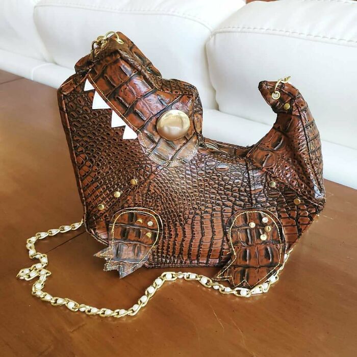Creative people’s cool crocodile-shaped leather handbag with unique eye detail and gold chain on wooden surface.