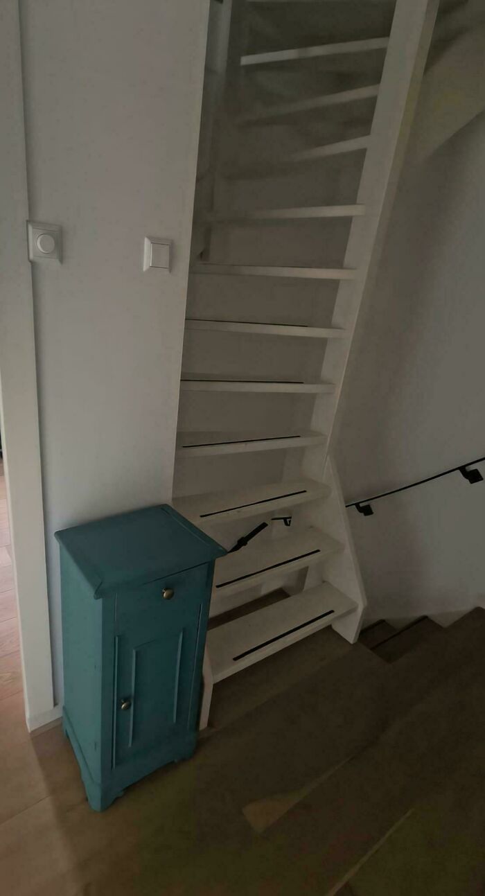 Narrow steep white stairs indoors with a small turquoise cabinet beside them in a dimly lit space.