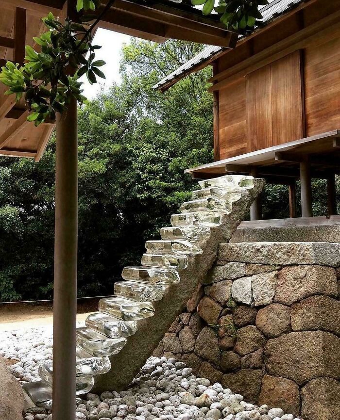 Unique transparent stairs leading to a wooden Airbnb structure, surrounded by stones and dense green forest foliage.