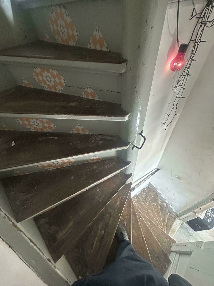 Steep narrow wooden stairs in a worn Airbnb rental with a red light bulb and patterned tiles on the walls.