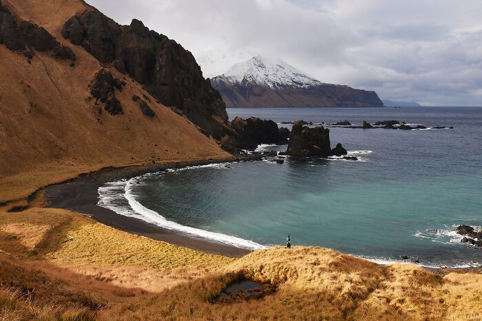 Remote location featuring rugged cliffs, black sand beach, and a snowy mountain backdrop by the ocean shore.