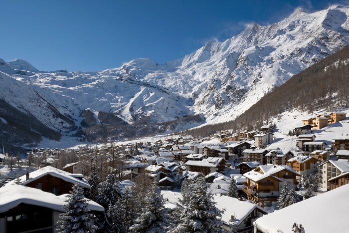 Snow-covered remote mountain village surrounded by towering peaks and clear blue sky in a stunning location.