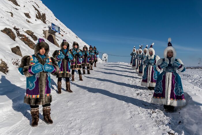 Group of people wearing traditional fur clothing standing on snowy terrain in a remote location with clear blue sky above.