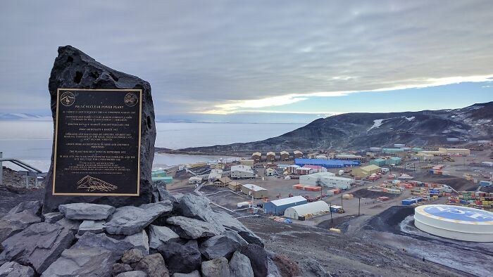 Plaque overlooking a remote icy research base on rocky terrain by the sea under a cloudy sky.