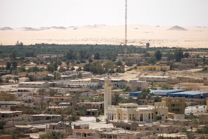 Remote desert location with buildings and a mosque surrounded by sparse vegetation and sand dunes in the background