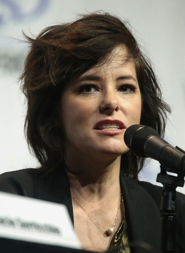 Parker Posey speaking into a microphone at an event, showcasing her career highlights and bio in a professional setting.