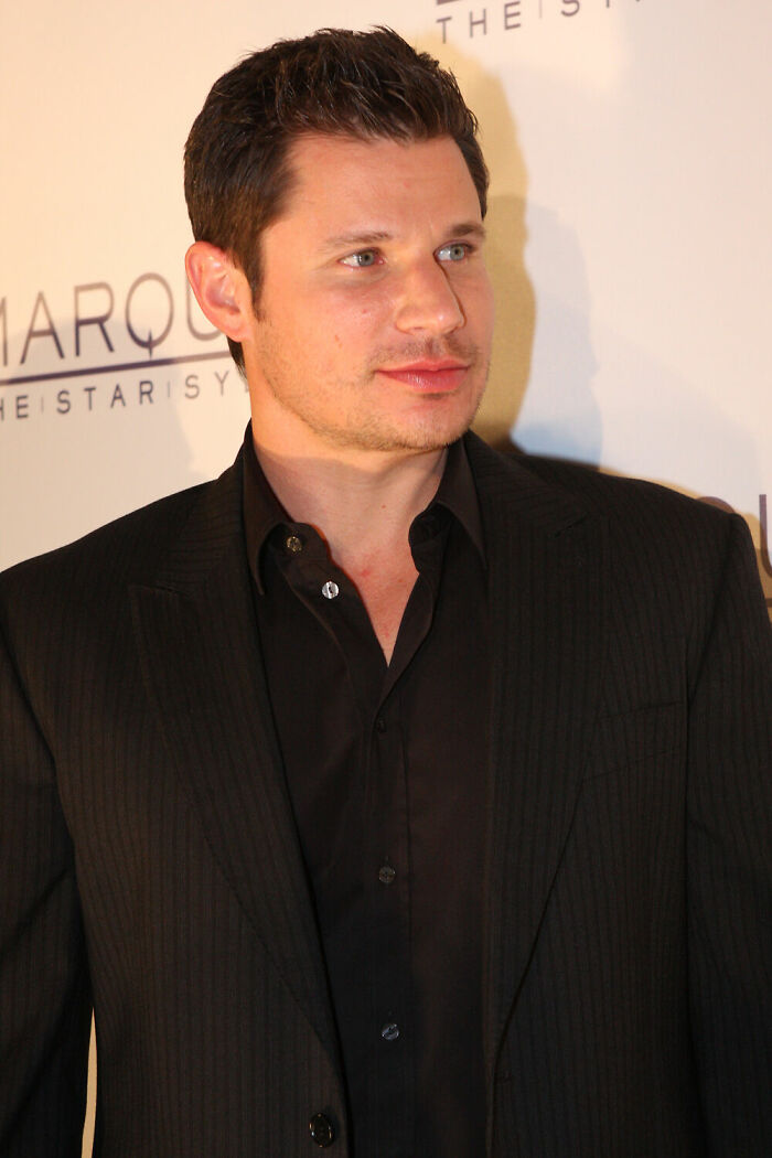 Nick Lachey wearing a black suit and shirt, posing at an event with a branded backdrop behind him.