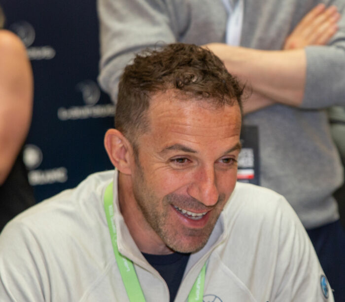 Alessandro Del Piero smiling and engaging during an event, wearing a white jacket and green lanyard.