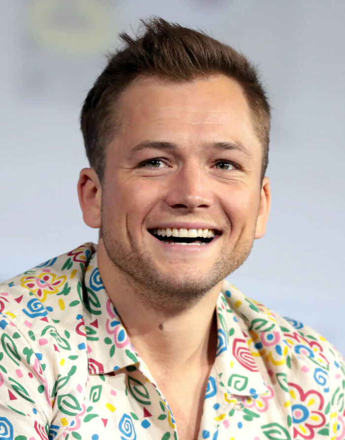Taron Egerton smiling and wearing a colorful patterned shirt during a public speaking or media event.