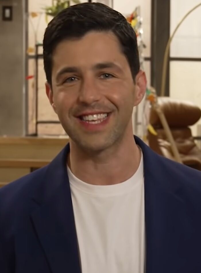 Josh Peck smiling indoors, wearing a white shirt and blue blazer, highlighting his bio and career achievements.