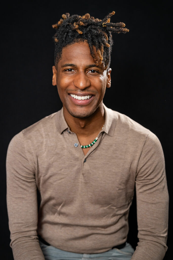 Jon Batiste smiling in a beige shirt with a necklace against a black background in a portrait photo.