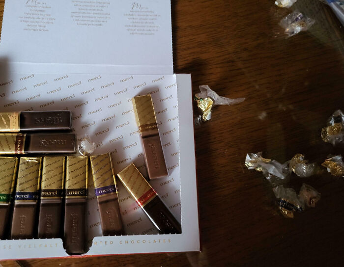 Partially eaten box of chocolates with several wrappers scattered on a wooden table, showing family frustration.