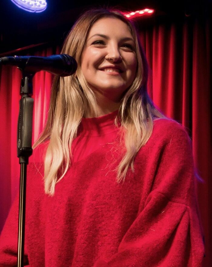 Julia Michaels smiling on stage near a microphone, wearing a red sweater, with red curtains in the background.