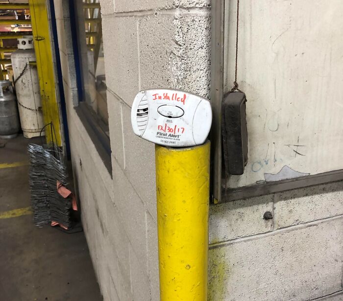 Smoke detector humorously installed on a yellow bollard in a warehouse, showing a clear example of people not even trying.