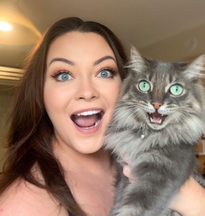 Woman with bright blue eyes holding a fluffy gray cat with green eyes, both appearing to take a better selfie than you.