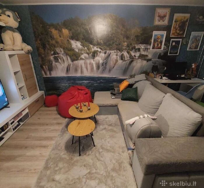 Cozy living room with waterfall mural, gray sectional sofa, red bean bags, and modern wooden nesting tables in real estate listing.