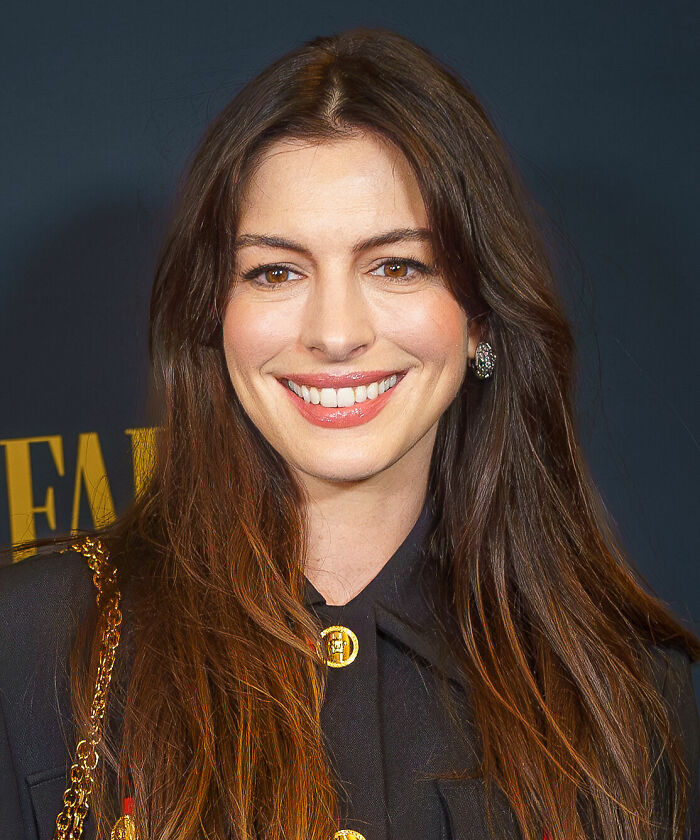 Anne Hathaway smiling with long brown hair, wearing a black outfit, at a formal event background.