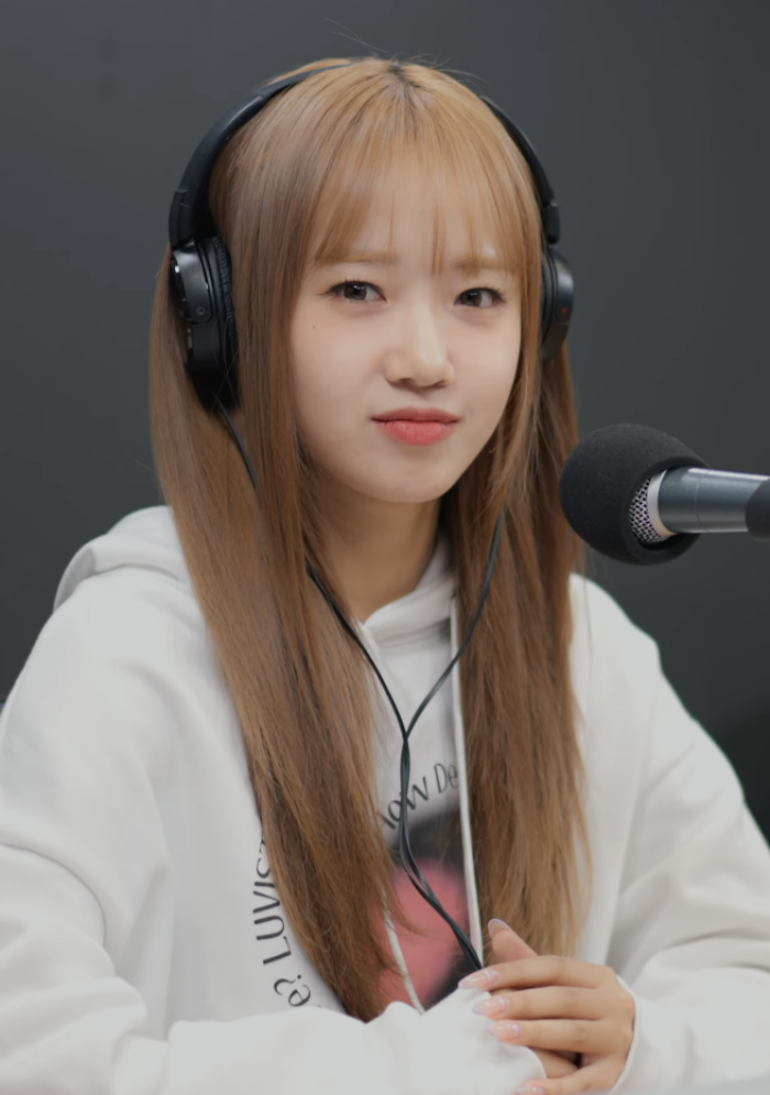 Choi Yoo-jung wearing headphones and a white hoodie, speaking into a microphone during a recording session.