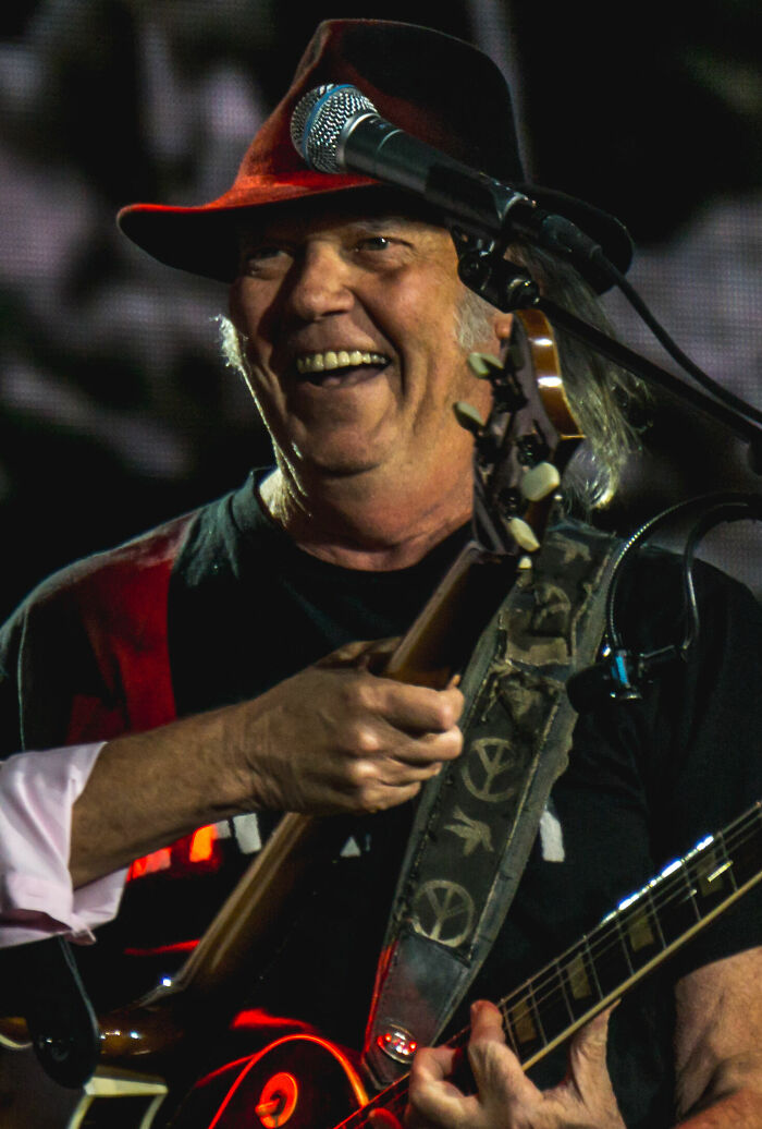 Neil Young smiling while playing guitar and singing on stage during a live music performance.