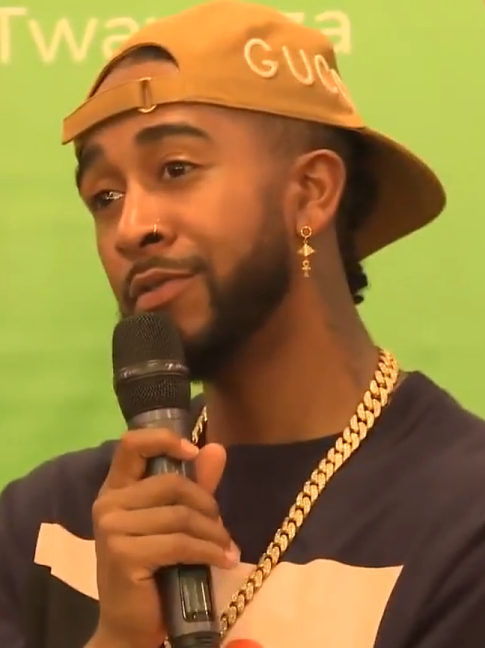 Omarion wearing a tan cap and gold chain, speaking into a microphone during a public event or interview session.