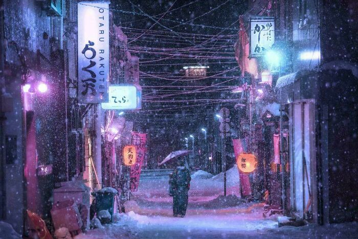 Snowy urban street at night with glowing signs and a person walking under an umbrella showing a different perspective.