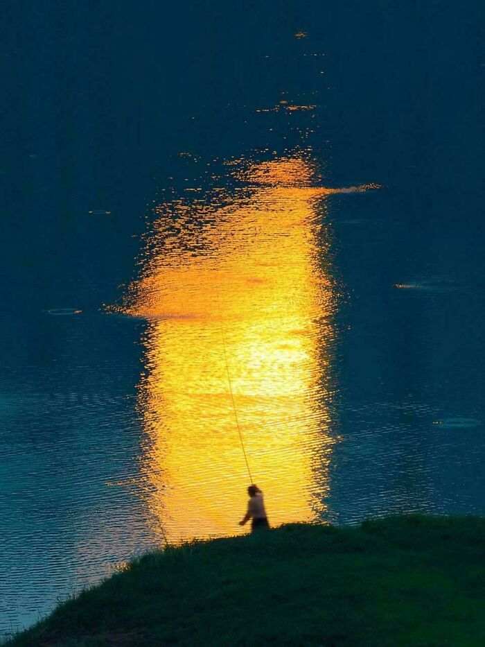 Silhouetted person fishing at sunset by water reflecting golden light, showing our world from a different perspective.