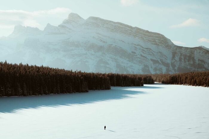 A lone person walks across a snowy landscape with towering mountains and dense forest, showing an amazing world perspective.