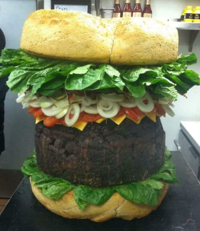 Giant burger with oversized patty, lettuce, onions, cheese, and bun stacked absurdly in a kitchen setting.
