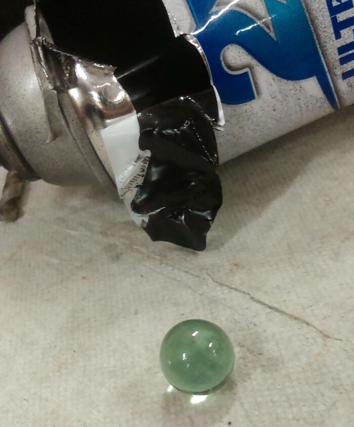 Close-up of the crushed spray can revealing its damaged interior and a small green marble on a textured surface.