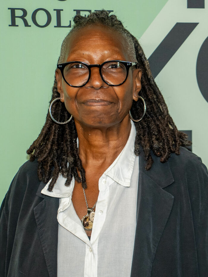 Whoopi Goldberg wearing glasses and hoop earrings at a public event, showcasing her bio and career highlights.