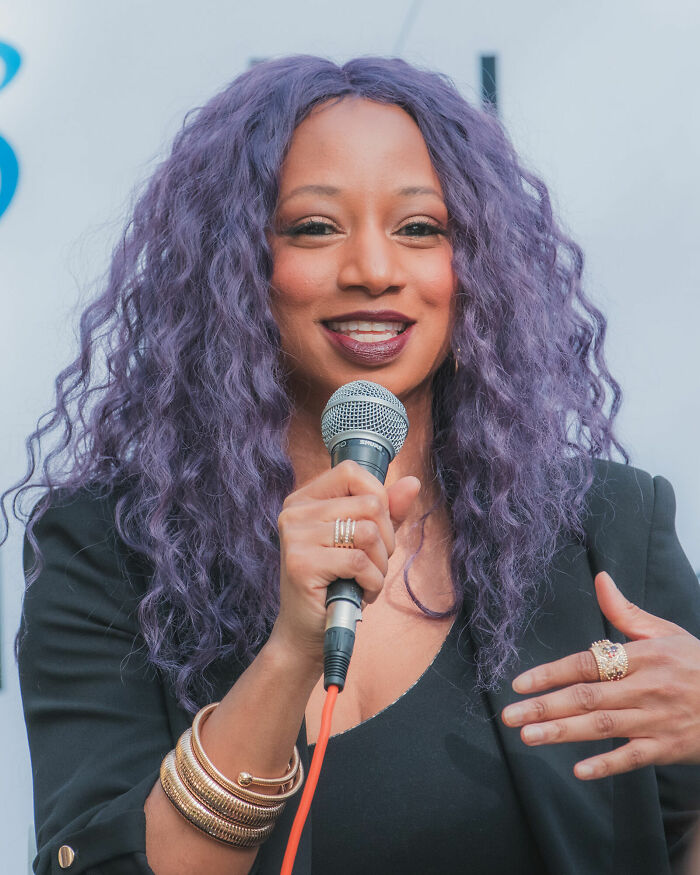 Monique Coleman speaking into a microphone, with purple curly hair and wearing black attire at an event.