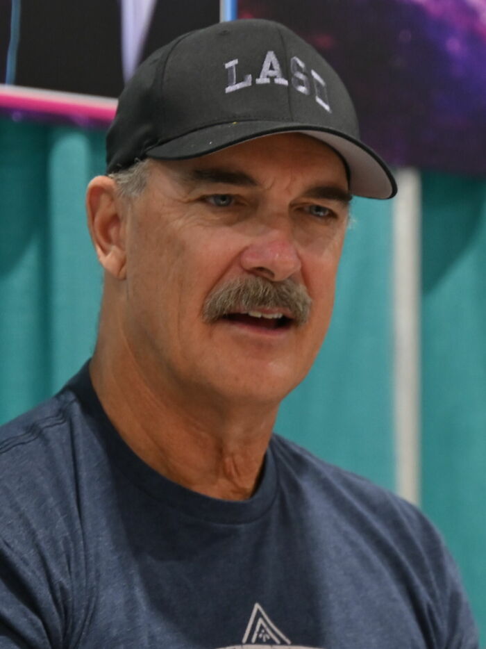 Patrick Warburton wearing a black cap and navy shirt, speaking at an indoor event with a turquoise backdrop.