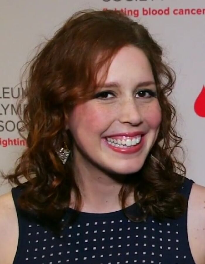 Vanessa Bayer smiling at an event, with curly red hair and wearing a navy blue dress with earrings.