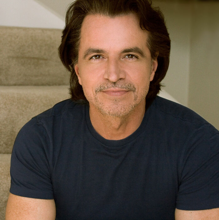 Yanni sitting on carpeted stairs, wearing a dark t-shirt, with a calm and confident expression, representing bio and career highlights.