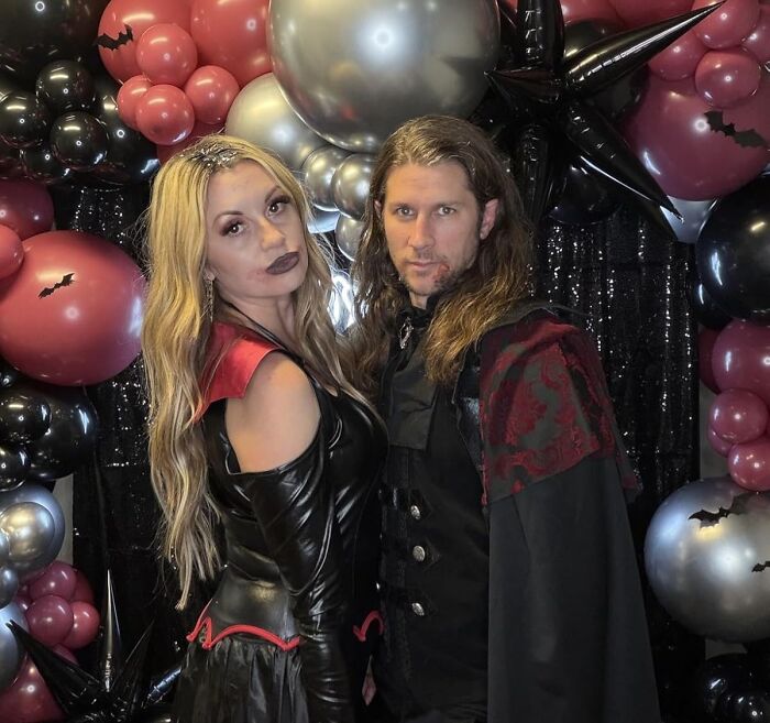 Couple dressed in Halloween costumes surrounded by festive black, red, and silver balloons for holiday shopping gifts ideas.