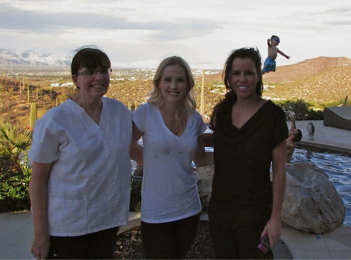 Three women smiling by a pool with a child photobombing mid-air jump in an epic photobombs moment outdoors.
