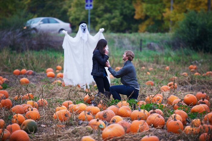 Couple in a pumpkin patch with a ghost photobombing their romantic proposal, capturing an epic photobomb moment.
