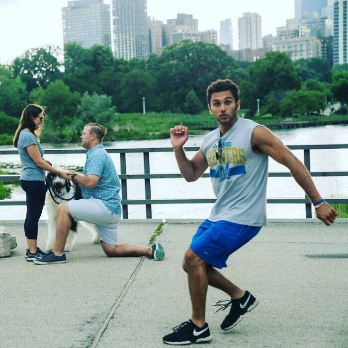 Man photobombing a couple's proposal by a river with city buildings and greenery in the background.