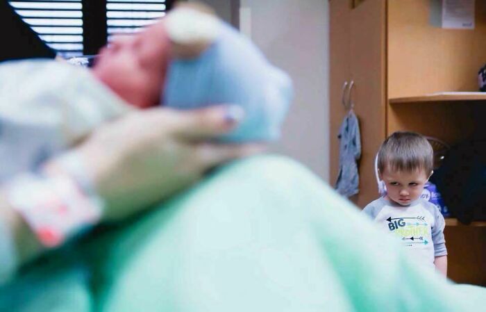 Toddler photobombing a newborn baby in a hospital room, creating an epic and funny moment with a mischievous look.