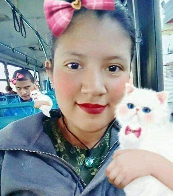 A woman holding a white cat with a bow, while a man photobombs the picture on a bus with a funny expression.