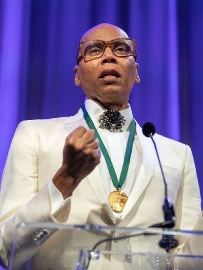 RuPaul speaking at an event, wearing a white suit and glasses with a medal around the neck.