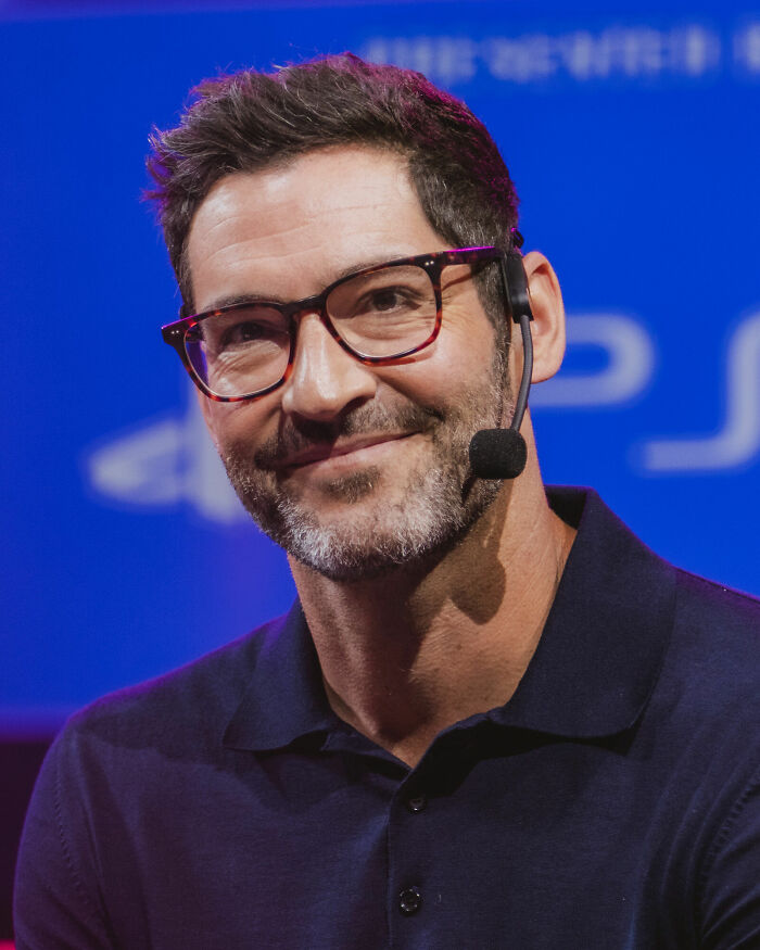 Tom Ellis wearing glasses and a headset microphone, smiling during a live event discussing his bio and career highlights.