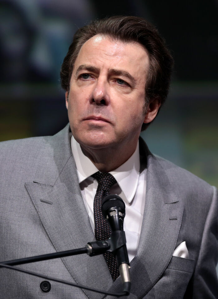 Jonathan Ross speaking into a microphone, dressed in a gray suit and patterned tie, during a formal event.