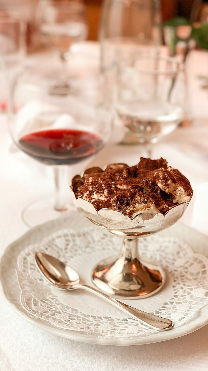 Dessert served in a silver dish with a spoon on a plate, next to a glass of red wine on a white tablecloth.