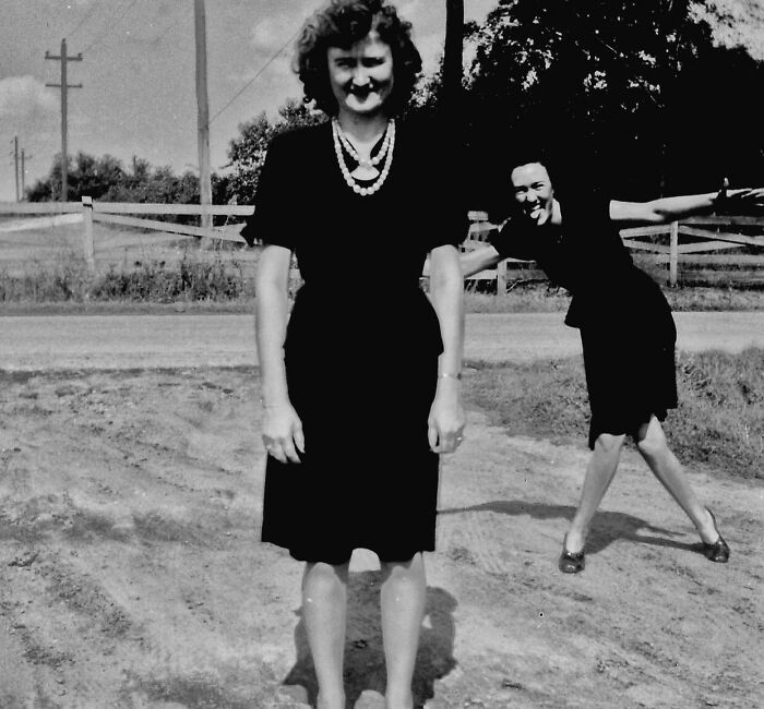 Two women in black dresses, one posing for a photo while the other photobombs with arms outstretched outdoors.
