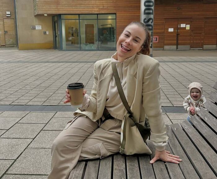 Woman holding coffee and smiling on bench with toddler photobombing and laughing in the background outdoors.