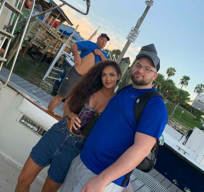 Couple posing on a dock with a man photobombing in the background lifting his shirt, an epic photobomb that stole the show.
