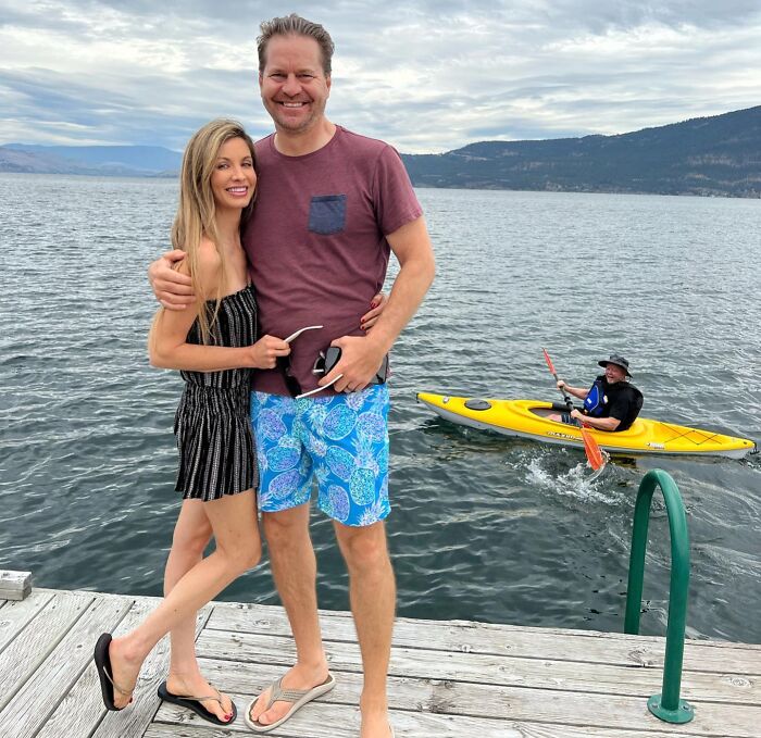 Couple posing on a dock with a man photobombing in a yellow kayak on the water behind them, capturing an epic photobomb moment.