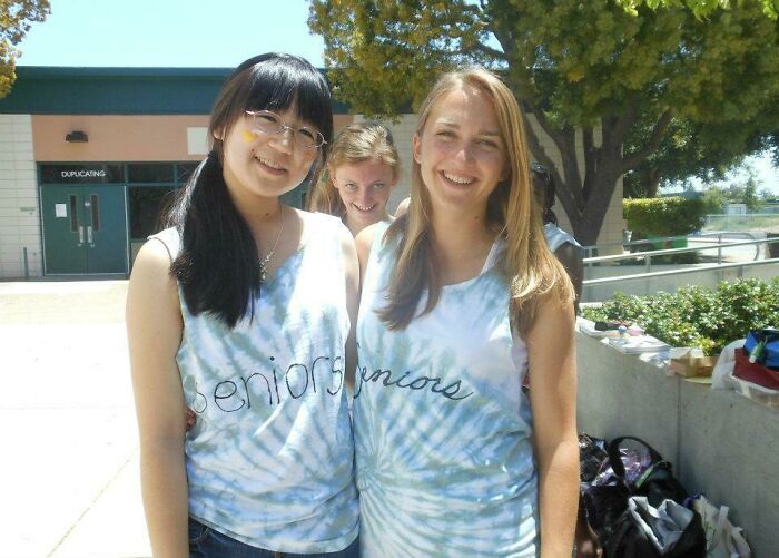 Two smiling girls wearing seniors tie-dye shirts with a photobomber making a funny face behind them outdoors.